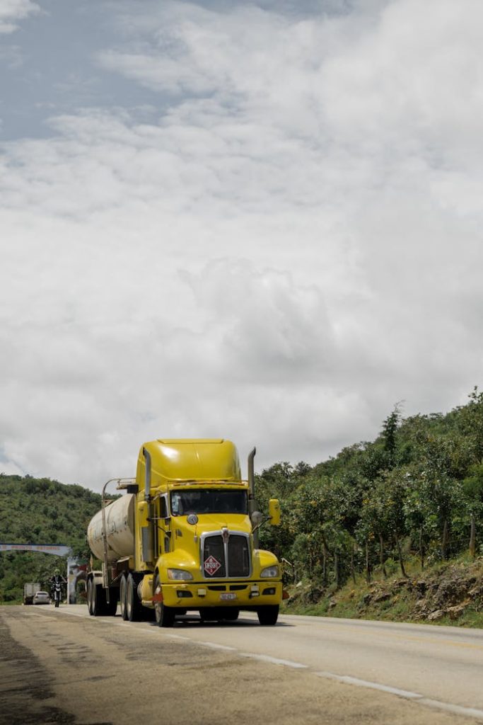 A yellow freight truck travels on a rural road under a cloudy sky.